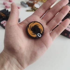 Pode incluir: Uma ficha de jogo circular preta e laranja com um design gráfico em espiral e um zero branco impresso na superfície. A ficha tem uma borda texturizada semelhante a uma engrenagem e um pequeno entalhe quadrado preto no centro do design.