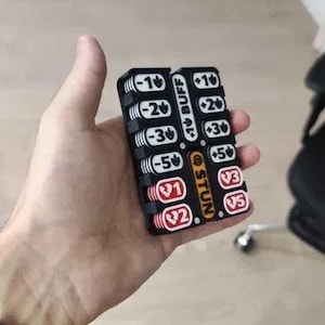 May include: A black plastic game tracker with two columns of sliding numbered tokens. The left column features white tokens with negative values and red tokens with heart icons. The right column has white tokens with positive values and red heart icons.