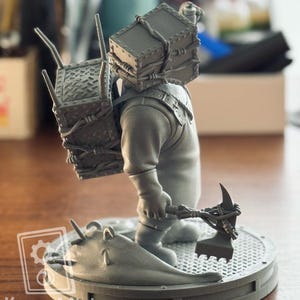 May include: A gray resin miniature figure standing on a circular base, wearing a large box-shaped headpiece and carrying a matching rectangular container on its back. The figure holds a spiked weapon and stands beside a slug-like creature.
