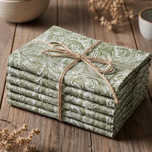 Pode incluir: Uma pilha de cinco guardanapos de tecido dobrados com um padrão paisley verde e branco. Os guardanapos estão cuidadosamente agrupados com um pedaço de corda castanha atado num laço à volta do centro da pilha, criando uma apresentação organizada.