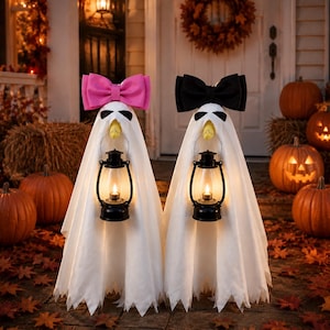 Puede incluir: Dos decoraciones de fantasmas blancos de pie uno al lado del otro. Cada fantasma presenta una cabeza puntiaguda con marcas oculares negras, un pico amarillo y una linterna negra al frente. Un fantasma lleva un lazo rosa, el otro un lazo negro.