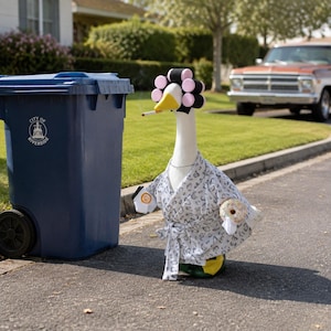 May include: A white goose figurine wearing a grey and white leopard print robe, pink and black hair rollers, and a thin silver necklace. The goose holds a coffee cup and a donut, with a cigarette in its beak, standing next to a blue trash bin.
