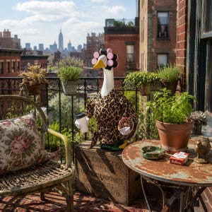 May include: A white goose figurine wearing a leopard print robe and pink hair rollers, holding a coffee cup and a chocolate donut. The goose has a cigarette in its beak and sits on a wooden crate labeled Garrett & Co. on a city balcony.