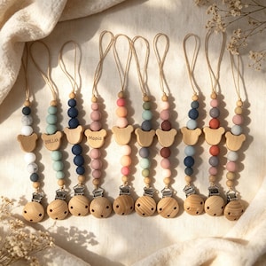 Puede incluir: Diez sujetachupetes dispuestos en fila, cada uno con un lazo de cuerda, una cuenta de madera con forma de oso, cuentas de silicona de varios colores y un clip de madera redondo. Dos clips tienen grabados los nombres Quillan y Mopia.