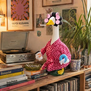 May include: A white goose figurine wearing a pink leopard print robe and pink hair rollers. The goose has a yellow beak, holds a white coffee cup and a blue donut, and has a cigarette in its beak. It stands on a green base.