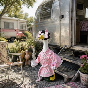 May include: A white goose figure wearing a pink and white heart-patterned robe and black hair rollers on its head. The goose holds a pink frosted donut in one wing and has a cigarette in its beak, standing outside a silver metal camper trailer.