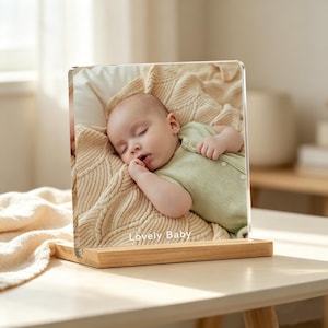 May include: A square acrylic photo block featuring a printed image of a sleeping baby wearing a light green onesie, resting on a cream-coloured textured blanket. The bottom of the acrylic panel displays the white text Lovely Baby.