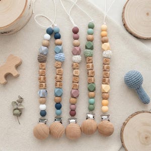 May include: Five pacifier clips featuring a mix of round and geometric silicone beads, crocheted spheres, and wooden cubes engraved with the word BABY. Each clip ends with a circular wooden fastener and is attached to a white cord loop for attachment.