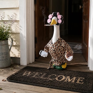 May include: A white goose figurine wearing a leopard print robe and pink hair rollers. The goose holds a white coffee cup and a chocolate donut, with a cigarette in its beak emitting a thin wisp of smoke, standing on a dark brown welcome mat.