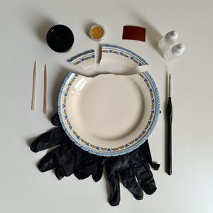 May include: A ceramic repair kit featuring a broken white plate with a blue and yellow floral rim, two wooden sticks, a black mixing cup, gold powder, a small rectangular sanding pad, two white adhesive bottles, a black paintbrush, and black gloves.