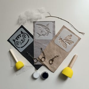 Op de afbeelding: Een knutselset met drie stoffen rechthoeken in zwart, grijs en beige, elk met een sjabloon van een kat, haai of bij. Bevat twee gele schuimkwasten, twee kleine ronde verfpotjes, drie metalen sleutelhangers, wit netmateriaal en een dun takje.