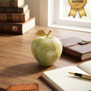May include: A decorative apple sculpture crafted from polished light green stone with natural veining. The fruit features a metallic gold-coloured stem and a single textured gold-coloured leaf attached to the top, creating a smooth, rounded appearance.