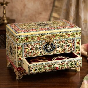 May include: A rectangular wooden jewellery box featuring intricate floral patterns in yellow, red, green, and blue. The box has a hinged lid with a gold-tone metal latch and a single pull-out drawer containing gold bangles and a pearl necklace.