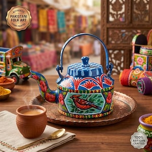 Pode incluir: Um bule de metal azul pintado à mão com uma ilustração de pássaro vermelho, padrões florais e uma tampa xadrez. O bule repousa sobre uma bandeja cor de cobre ao lado de uma pequena xícara de barro com chá, uma colher dourada e um guardanapo, com um emblema AUTHENTIC PAKISTANI FOLK ART HAND-PAINTED.