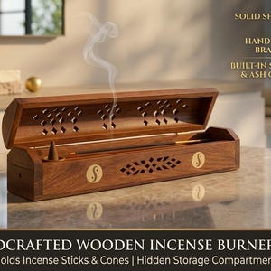 May include: A rectangular wooden incense burner box with a hinged lid, featuring brass inlay designs including yin-yang symbols and geometric cutouts. The box holds a burning incense cone and stick, with text stating solid sheesham wood.