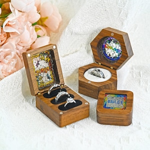 Puede incluir: Tres cajas de madera para anillos con tapas abatibles. Una caja rectangular contiene tres anillos de plata, una caja hexagonal contiene un anillo de plata y una pequeña caja cuadrada está cerrada. Cada tapa presenta una incrustación de mosaico.