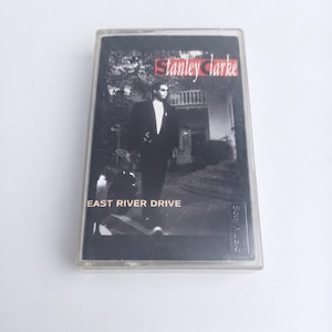 Puede incluir: Un casete del álbum East River Drive de Stanley Clarke. La portada presenta una fotografía en blanco y negro de una persona con traje frente a un edificio. El texto Stanley Clarke y East River Drive está impreso en la parte frontal.