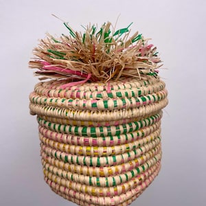 May include: A cylindrical woven basket with a lid, constructed from natural tan fibers interspersed with vibrant pink, green, and yellow accents. The top of the lid features a decorative, upright tuft of frayed, multi-colored raffia strands.