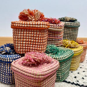 May include: A collection of square woven baskets with lids, each featuring a distinct color including orange, pink, blue, green, and yellow. Every basket is topped with a decorative raffia pom-pom in a matching or complementary shade.