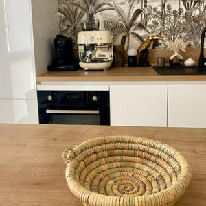 Peut inclure: Un panier rond et peu profond tressé à partir d'herbes séchées ou de paille de couleur claire. Le panier présente une construction en spirale avec un bord épais et renforcé ainsi qu'une petite anse sur le côté, offrant un aspect organique.