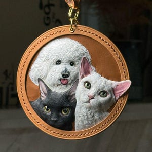 Peut inclure: Un porte-clés circulaire en cuir beige présentant un relief détaillé et peint de trois animaux : un chien blanc au pelage duveteux, un chat noir aux yeux jaunes et un chat blanc aux oreilles teintées de rose. Le cuir est cousu sur le bord.