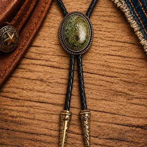 May include: A bolo tie featuring an oval-shaped green stone set in a twisted bronze-toned metal frame. The tie consists of a black braided cord with two matching bronze-toned metal tips at the ends, each decorated with intricate engraved patterns.