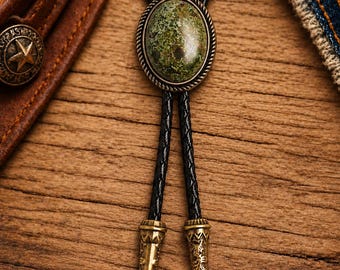 Puede incluir: Una corbata bolo con una piedra ovalada de color verde engastada en un marco de metal trenzado en tono bronce. La corbata consta de un cordón trenzado negro con dos puntas de metal a juego en tono bronce, decoradas con grabados intrincados.