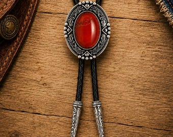 Puede incluir: Una corbata bolo con una piedra roja ovalada central engastada en un marco de metal plateado adornado con grabados intrincados. El colgante está unido a un cordón trenzado negro que termina en dos puntas decorativas de metal plateado a juego.