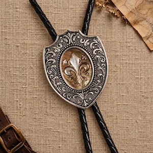 May include: A bolo tie featuring a silver-toned metal shield-shaped slide with intricate scrollwork and a central oval inlay of iridescent mother-of-pearl. The inlay displays a raised fleur-de-lis emblem. It includes a black braided cord.