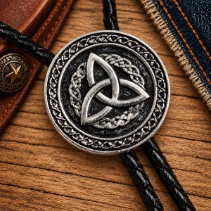 May include: A bolo tie featuring a circular silver-toned pendant with a raised triquetra knot symbol at the center, surrounded by an intricate braided border. The pendant is attached to a black braided cord that hangs down from the circular piece.