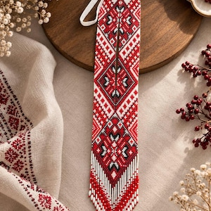 Puede incluir: Un collar de cuentas largo y estrecho con un patrón geométrico en rojo, blanco y negro. El diseño consta de intrincados motivos de diamantes y cruces, que terminan en un fleco puntiagudo de cuentas rojas y blancas con un lazo de cordón blanco.