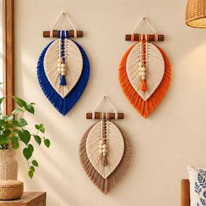 Pode incluir: Três tapeçarias de macramé em forma de folha, cada uma pendurada num bastão de madeira escura. Os designs apresentam cordão cor de creme em camadas com franjas azuis, laranja ou castanhas, acentuadas por três contas de madeira clara.