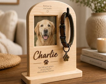 May include: A light-colored wooden memorial frame with an arched top, featuring a photo of a golden retriever, a black hook holding a green dog collar with a paw print charm, and the engraved text "Forever in our hearts", "Charlie", and "You left paw prints on our hearts that will last forever."