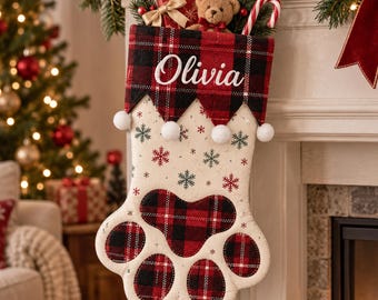 May include: A paw-shaped Christmas stocking featuring a cream-colored body with red and green snowflake patterns. The cuff and paw pads are made of red and black plaid fabric. The name Olivia is embroidered in white script on the cuff.