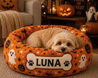 May include: A round orange pet bed featuring a Halloween pattern of black bats, white ghosts, and jack-o'-lanterns. A central white bone-shaped patch on the front displays the name LUNA in black letters, flanked by two black paw print icons.