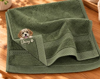 May include: A folded olive green cotton towel featuring an embroidered illustration of a light brown puppy with floppy ears and a small white paw print icon above the name Daisy embroidered in white cursive script lettering.