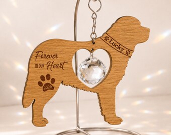 May include: A wooden dog-shaped ornament hanging from a silver metal stand. The wood features the engraved text "Forever in our Heart", "Lucky", and "2026" with a paw print. A clear faceted crystal sphere hangs inside a heart-shaped cutout.