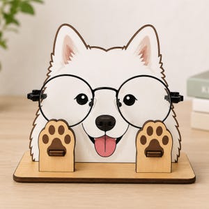 May include: A wooden eyeglass holder shaped like a white dog with pointed ears, a black nose, and a pink tongue. The dog has two raised paws that serve as supports for a pair of round, black-rimmed glasses resting across its face.