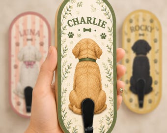 May include: An oval-shaped wooden wall hook featuring a golden retriever illustration, the name CHARLIE, paw prints, and a bone icon. The design includes green leaf accents and a black metal hook at the bottom. The background is cream-colored.