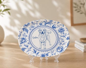May include: An oval ceramic plate featuring a blue and white floral pattern. The center displays a detailed illustration of a dog standing inside a circular border, with the name Sailor written in cursive script to the left of the dog.