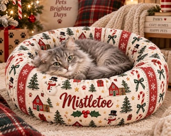 May include: A round pet bed with a cream-colored fabric featuring a Christmas pattern of snowmen, pine trees, red houses, and candy canes. The word Mistletoe is printed in red script on the front. A gray tabby cat rests inside the bed.