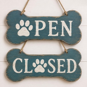 Op de afbeelding: Twee botvormige houten bordjes met een verweerde blauwgroene afwerking en witte letters. Het bovenste bordje toont een pootafdruk en het woord OPEN, terwijl het onderste bordje een pootafdruk en het woord CLOSED bevat. Beide hangen aan jutekoord.