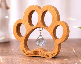 May include: A light brown wooden paw print ornament featuring a faceted clear crystal hanging from the center. The wood is engraved with the name BINGO, two leafy branch icons, and the years 2019 and 2026 on the bottom curved edge.