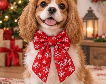 May include: A Cavalier King Charles Spaniel wearing a red fabric bow tie with a white snowflake pattern. The bow tie features two long, pointed tails, one of which is embroidered with the name Belly in white cursive script lettering.