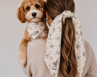May include: A cream-colored dog bandana with a blue and brown floral pattern featuring the name Daisy in black script, and a matching hair scrunchie with long fabric tails featuring the name Bella in black script, both made of matching fabric.