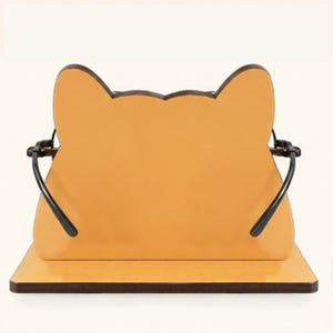 May include: A light orange, cat-shaped eyeglass holder featuring a flat base and a vertical panel with pointed ears. The stand includes two black metal arms extending from the sides to support the frame of a pair of glasses in a resting position.