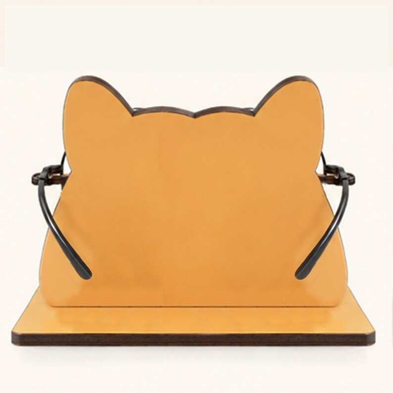 May include: A light orange, cat-shaped eyeglass holder featuring a flat base and a vertical panel with pointed ears. The stand includes two black metal arms extending from the sides to support the frame of a pair of glasses in a resting position.