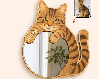 May include: A circular wall mirror framed by a wooden cutout of a brown tabby cat. The cat has green eyes, striped fur, and a long tail. The name Daisy is engraved on the cat's left paw. A small inset photo of the cat is shown in the corner.