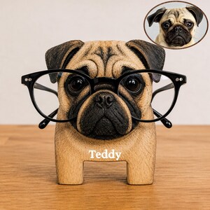 May include: A tan and black pug-shaped eyeglass holder with a black nose and ears. The figure features the name Teddy printed in white on the front. A pair of black-rimmed glasses rests on the pug's face. A small circular inset shows a real pug.