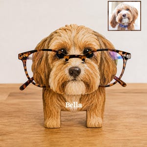 May include: A brown dog-shaped eyeglass holder with textured fur details, featuring the name Bella printed in white on the chest. The figurine is wearing a pair of brown tortoiseshell-patterned glasses across its face.